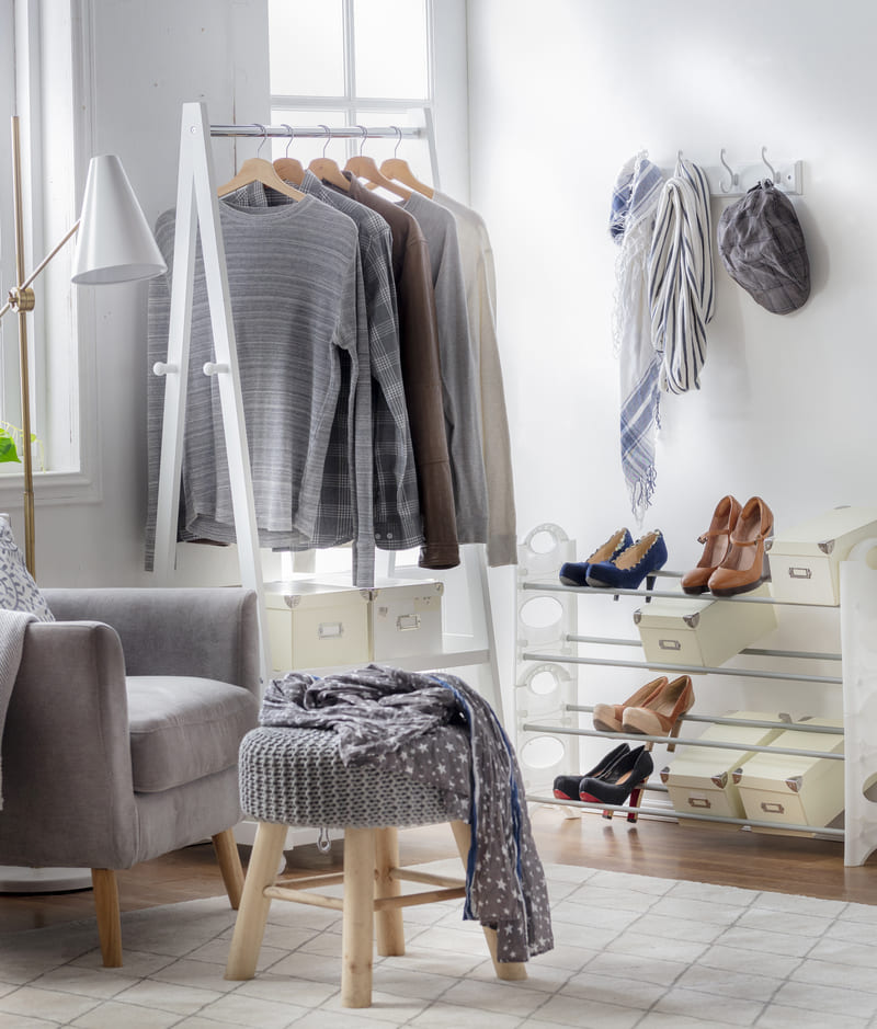 Espacio del hogar con una poltrona gris, un pouf, zapatero con varios zapatos y cajas, un perchero blanco con prendas colgadas, una lámpara de pie con pantalla blanca y base dorada y una alfombra blanca.