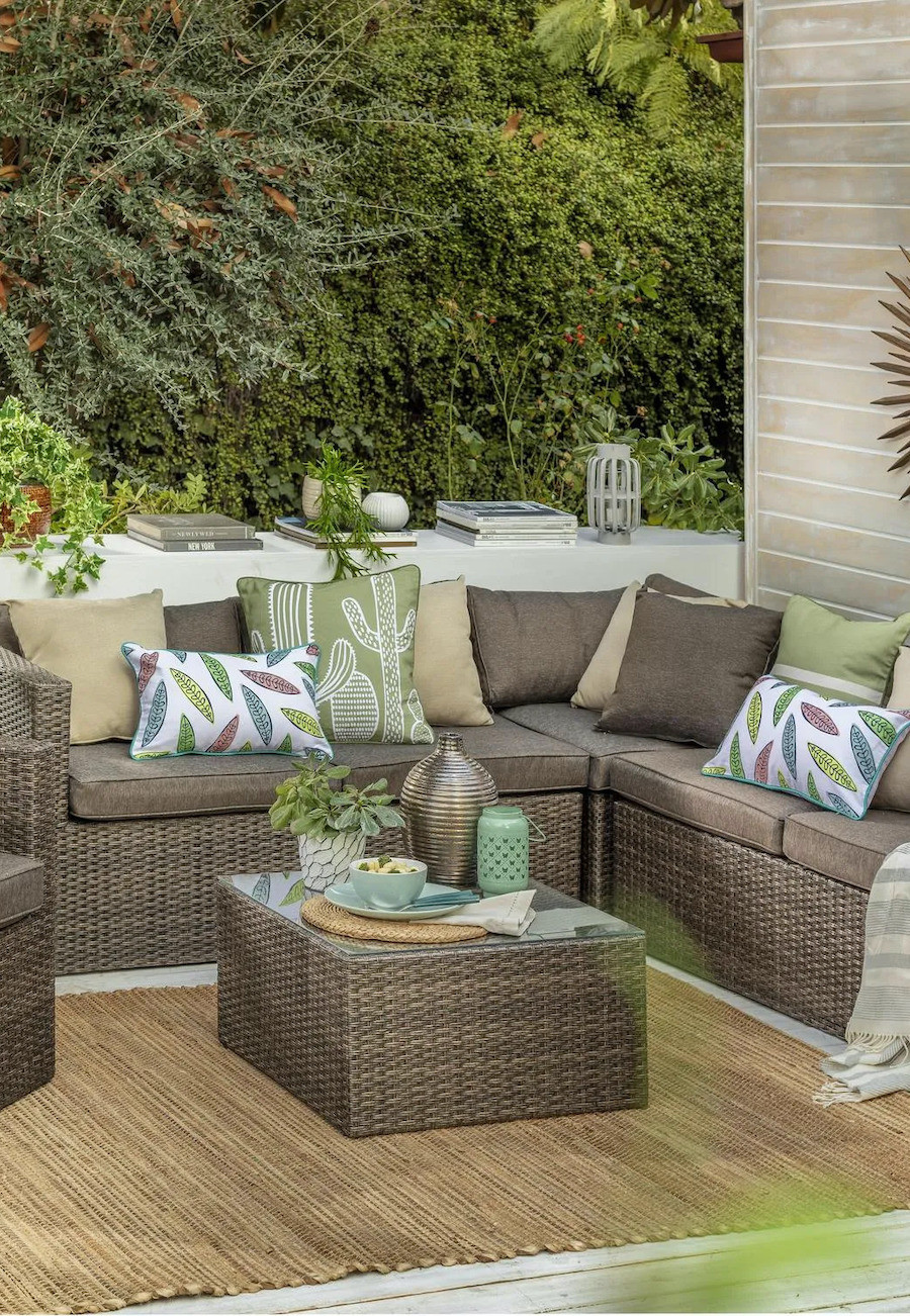 Terraza con muebles de ratán con cojines café, verdes y beige, y otros con hojas en tonos pastel. En el suelo hay una gran alfombra de yute y alrededor hay mucha vegetación.