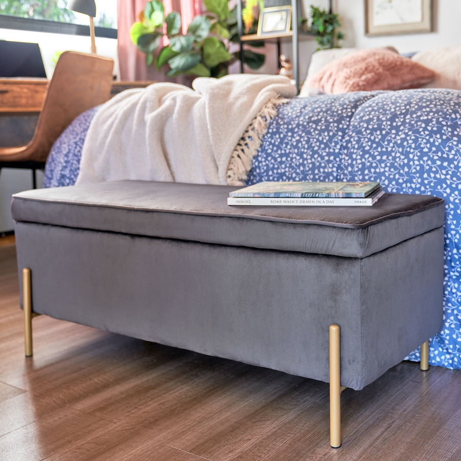 Banqueta multifuncional, con baúl escondido, de color gris y patas doradas. Esta puesta a los pies de una cama que tiene un plumón azul y una manta beige.