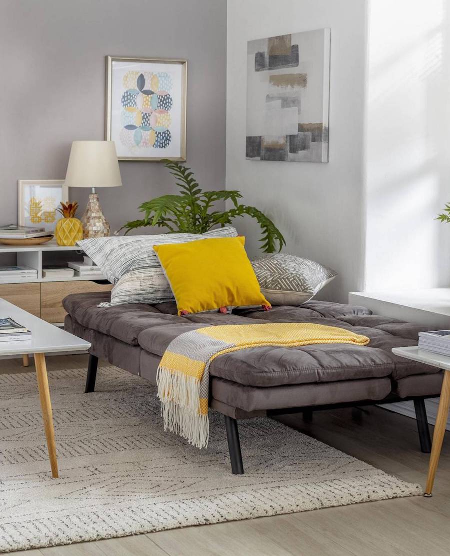 Futón abierto en un dormitorio. Es de color gris oscuro y sobre él hay almohadas, cojines y una manta. En la parte de atrás hay un mueble de madera con una lámpara, adornos y un cuadro.