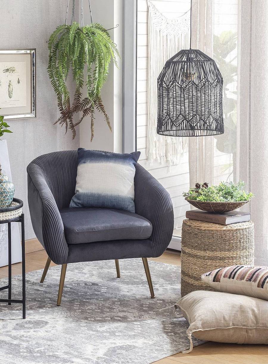 Pouf redondo multifuncional, que además sirve como mesa de apoyo. Es de mimbre en tonos café oscuro y claro. Arriba tiene un libro y una planta. Está al costado de una poltrona gris con patas de madera