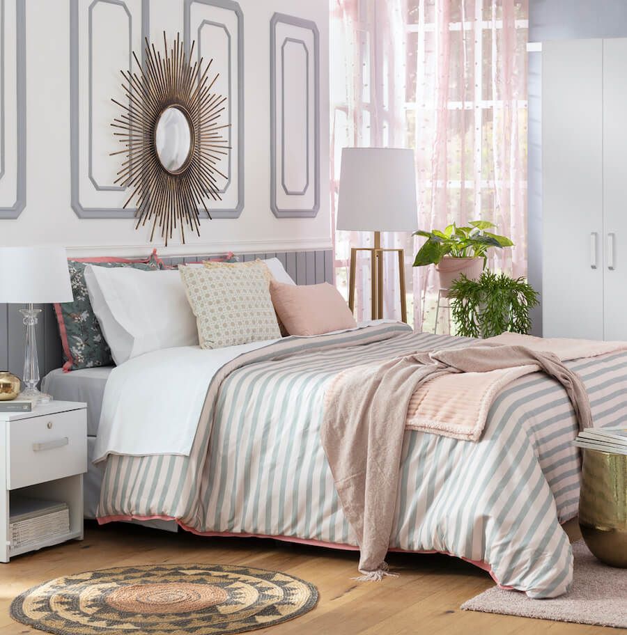 Dormitorio con decoración en tonos suaves, cama con ropa de cama a rayas en colores pastel, cojines , lámpara de pie junto a la ventana con visillos rosas, espejo decorativo en la pared, y plantas verdes en macetas.