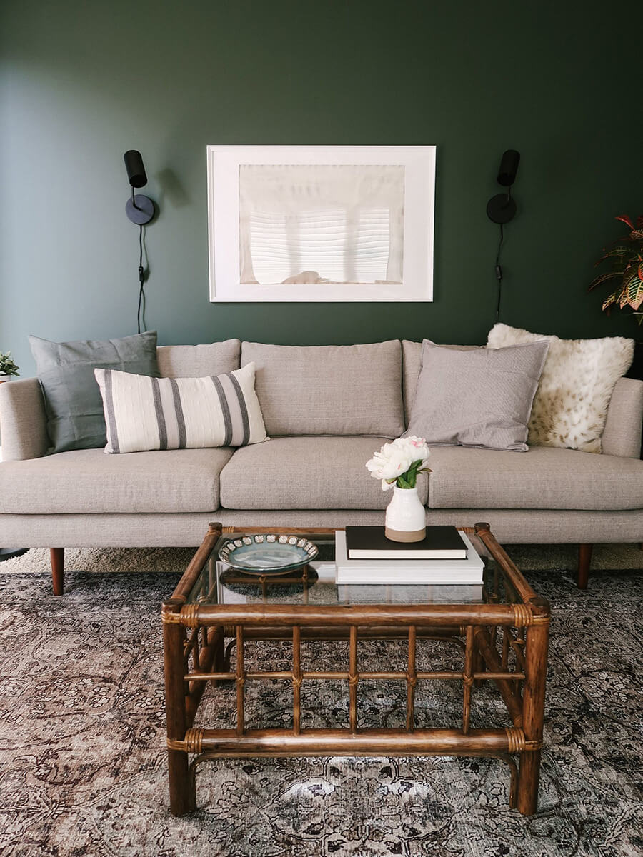 un living con un gran sofá de 3 cuerpos de color beige, con una mesa de centro de vidrio y madera, y las paredes pintadas con esmalte al agua color verde oscuro.