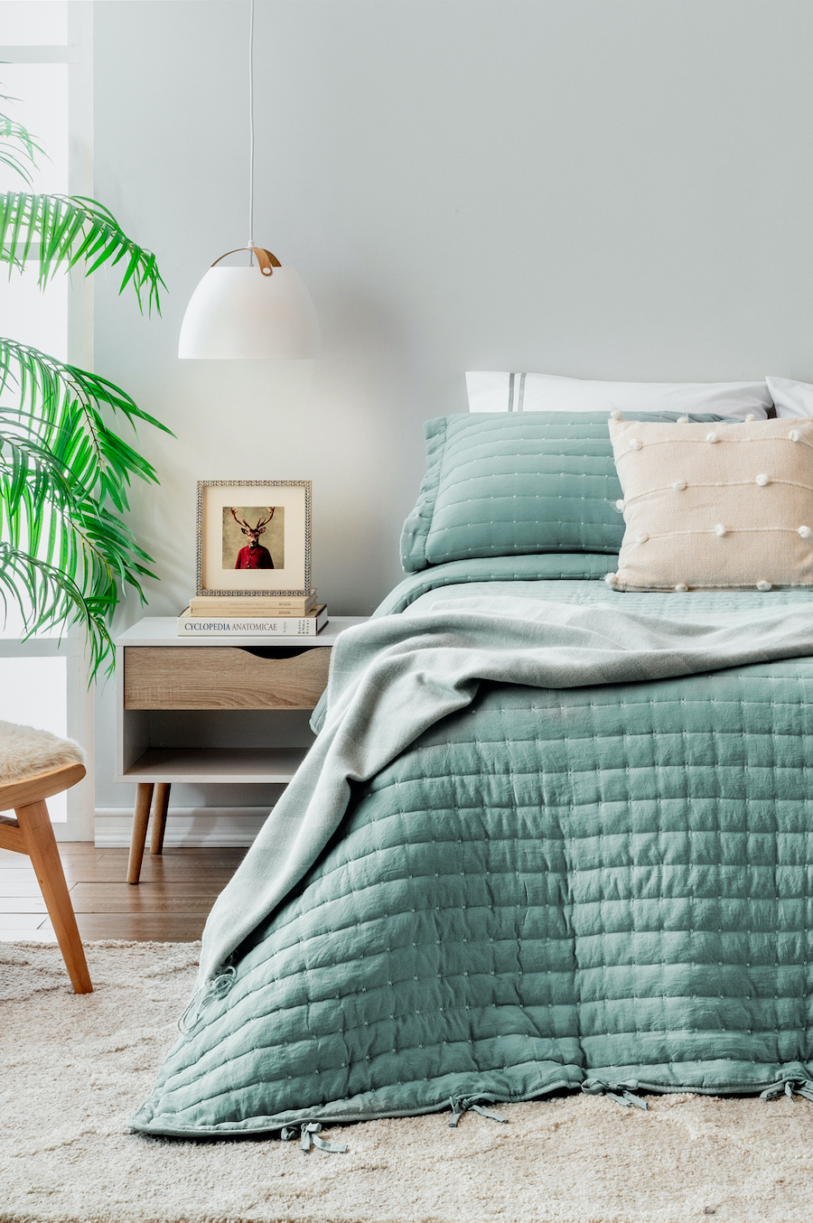 Dormitorio clásico con cama con cobertor color verde matcha y cojín beige. Velador con cajón y lámpara colgante blanca.