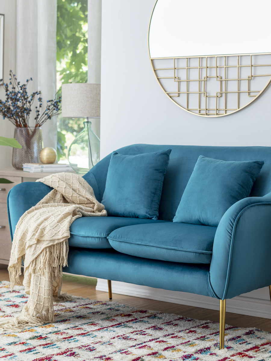 Living estilo clásico con sillón azul moderno, de dos cuerpos. Sobre él hay una manta beige. Muros blancos con espejo dorado y diseño de reja, Atrás se ve una mesa con lámpara de vidrio y pantalla beige y gran jarrón de vidrio con ramas.