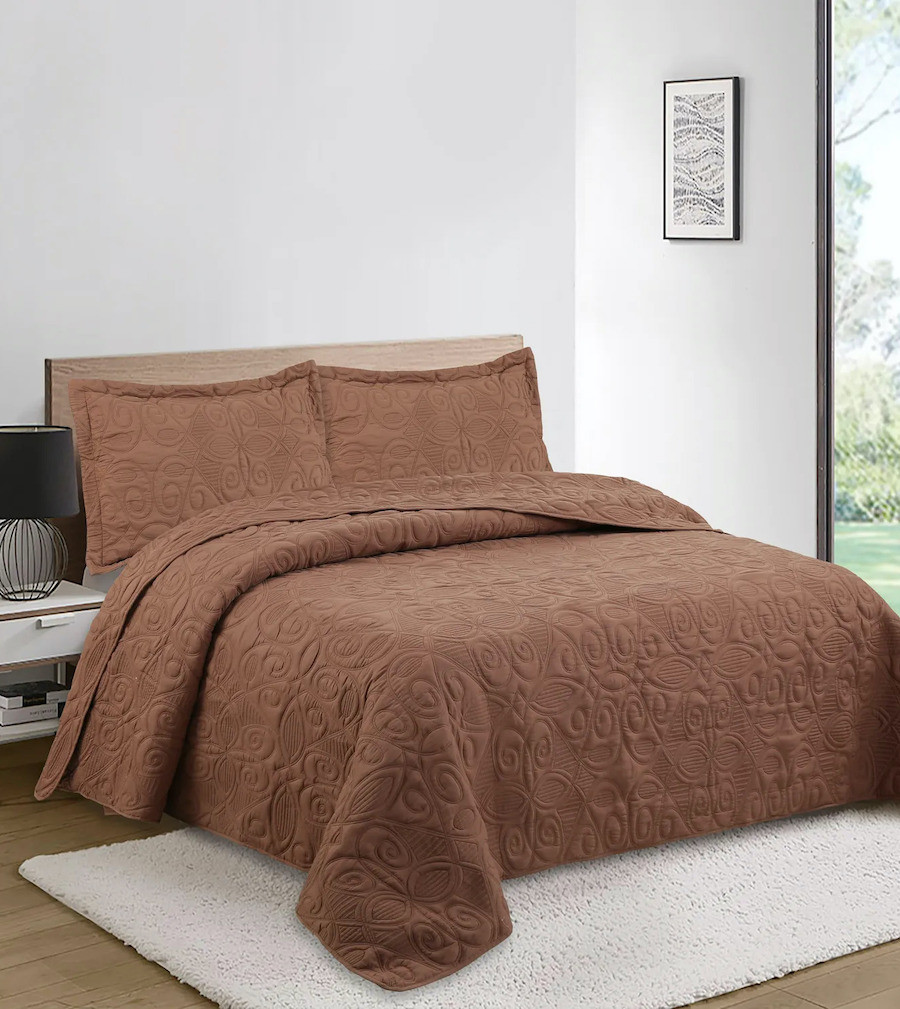 Dormitorio de paredes blancos con una cama de 2 plazas con un quilt marrón oscuro. En el piso hay una alfombra blanca.