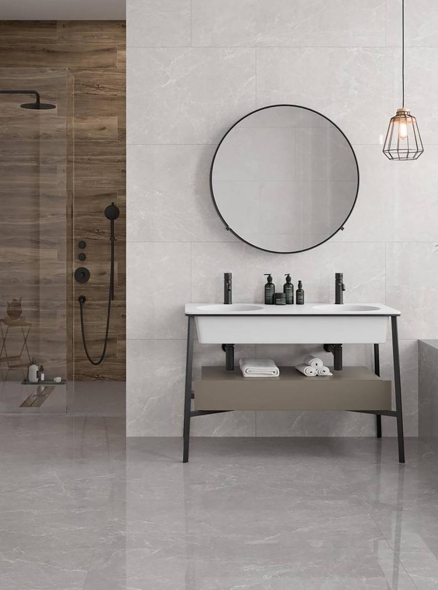 Baño principal con muros y pisos de porcelanato imitación mármol. Hay un gran espejo redondo con marco negro, un mueble vanitorio con patas negras, grifería negra y lavamanos blanco. Al fondo hay una ducha con muros de cerámica imitación madera.