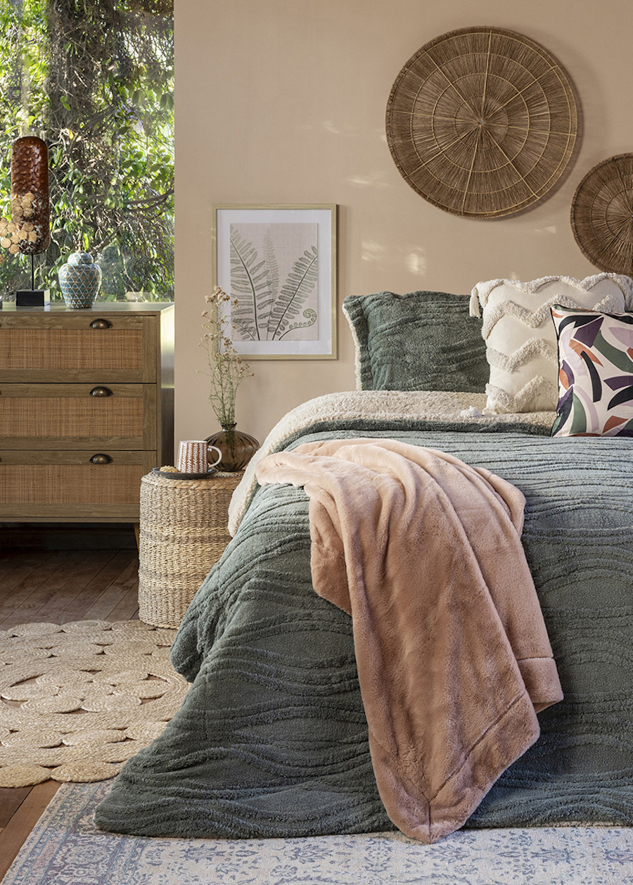 Pieza matrimonial decorada al estilo Boho chic con una cama con quilt verde, cojines y una manta rosa palo. En la pared de color beige hay platos de mimbre. Alrededor de la cama hay una alfombra de yute, una cómoda de madera y ratán.