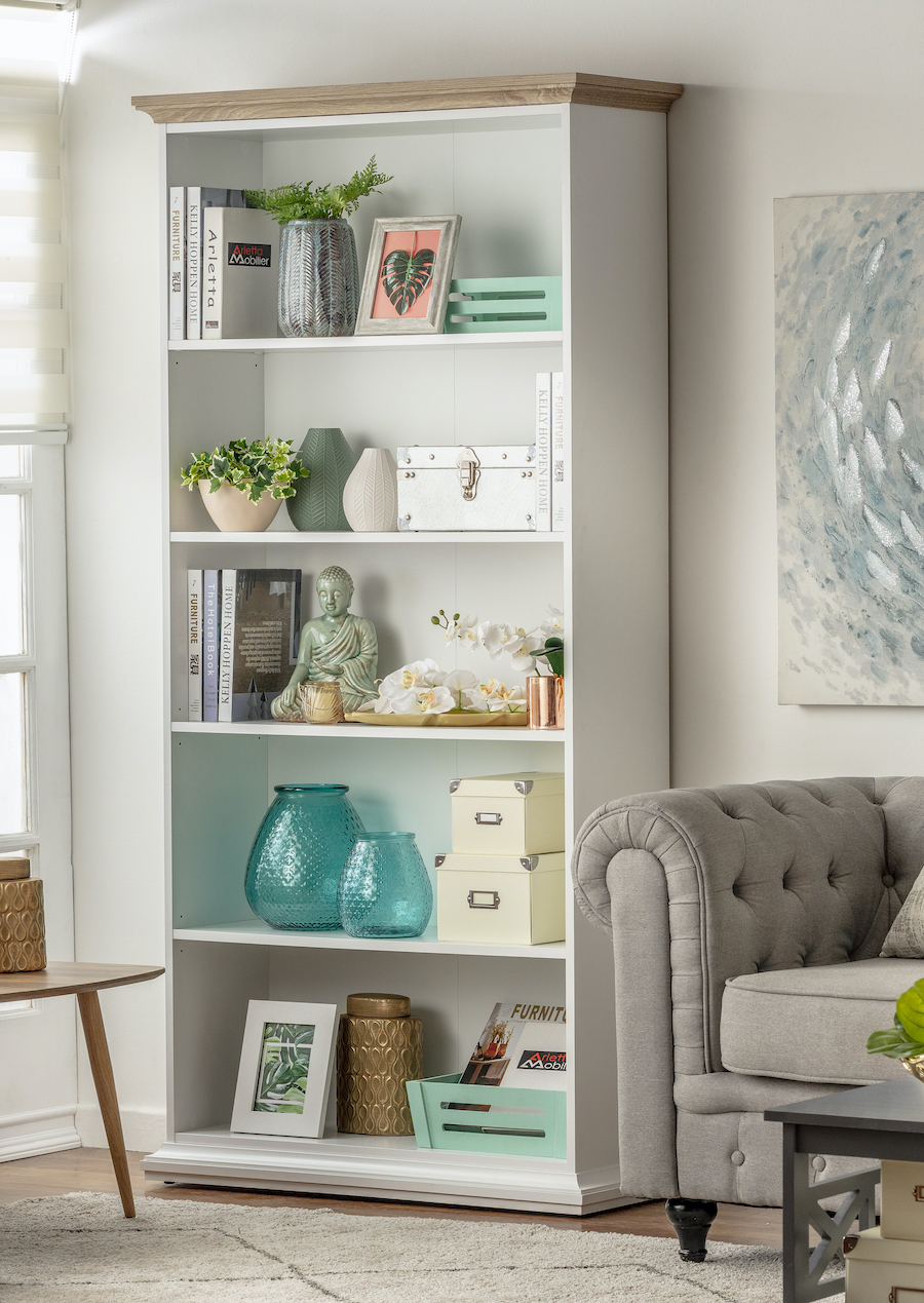 Gran estante vertical en living clásico. Es blanco y tiene 5 niveles con libros y adornos. Está junto a un sofá gris de estilo capitoné.