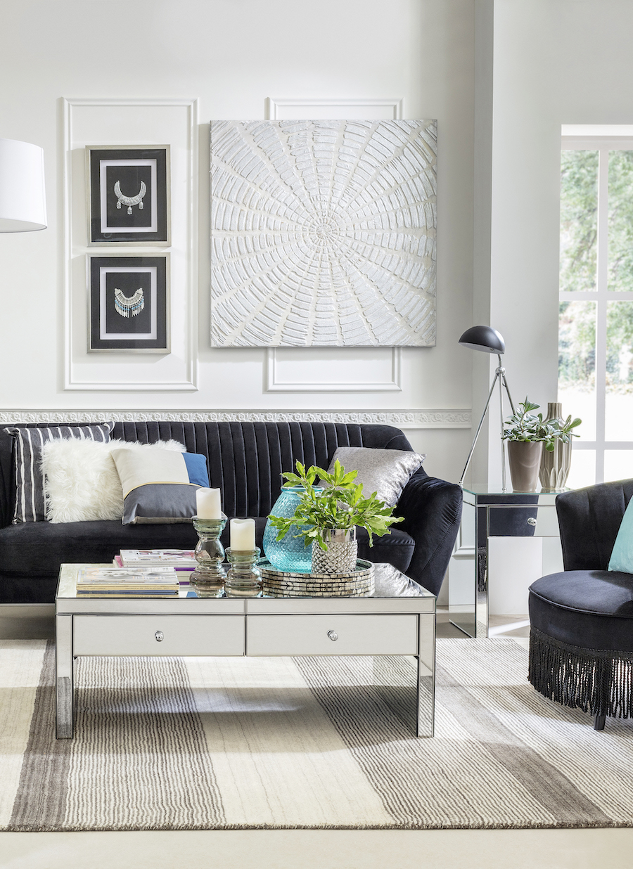 Living de estilo moderno con mesa de centro espejada, baja y con dos cajones. Sobre ella hay plantas, velas, libros y adornos. Sofá negro con cojines blancos, negros y plateados.