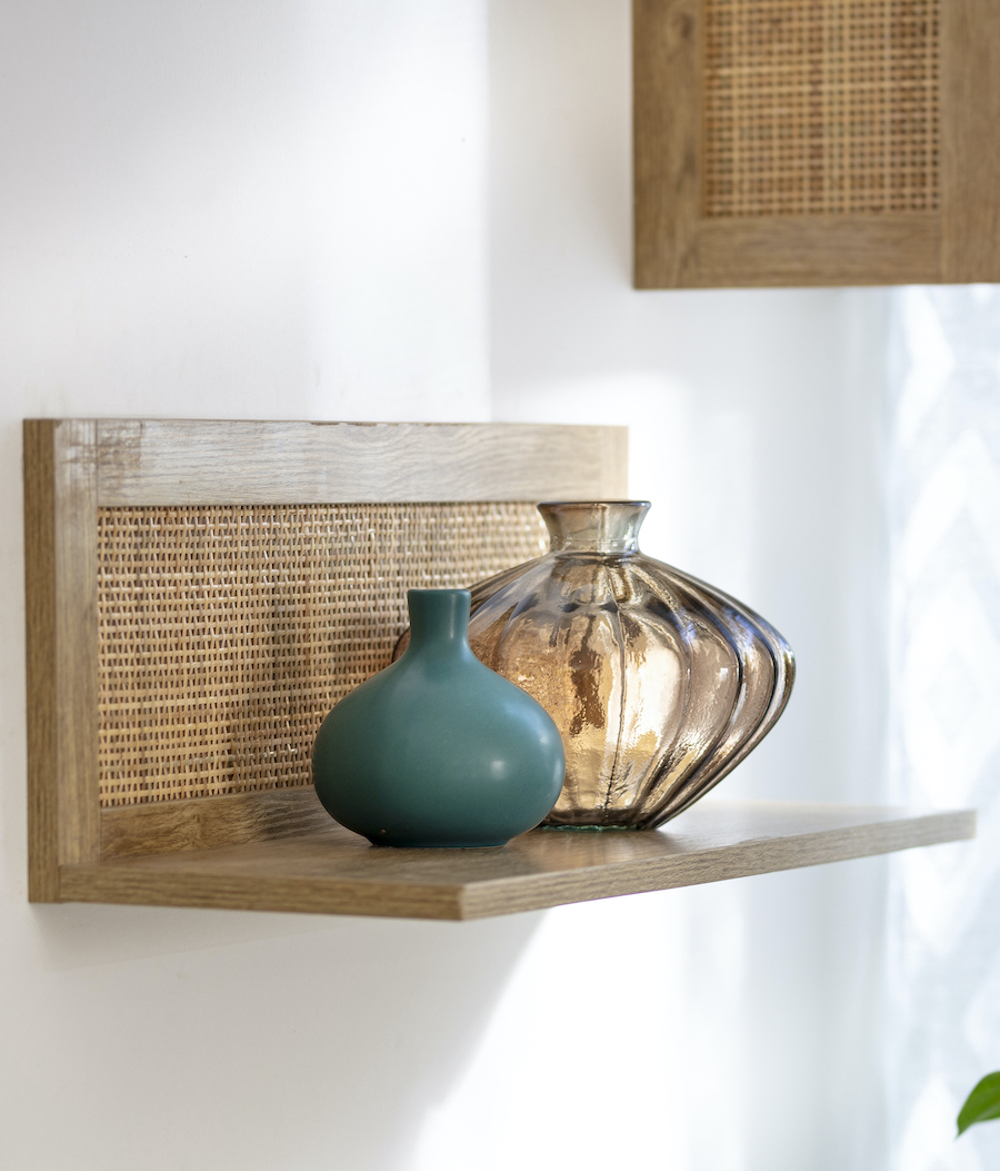Detalle de muro blanco con una repisa flotante de madera con junquillo. Sobre ella hay una botella transparente y un jarrón azul