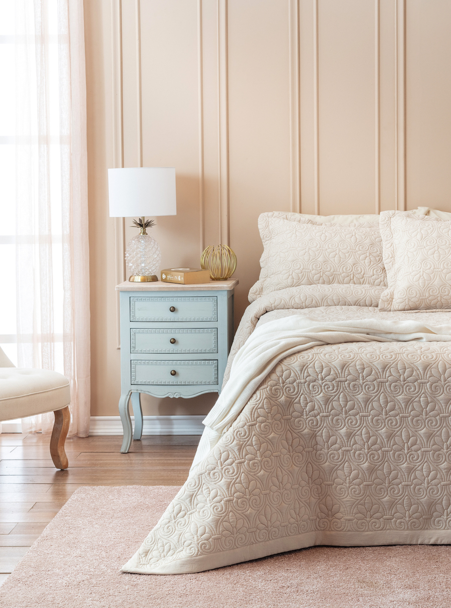 Dormitorio clásico con una cama con colcha acolchada de color beige y almohadas blancas. Junto a la cama hay un velador celeste con tres cajones, con una lámpara blanca con forma de piña y un elemento decorativo