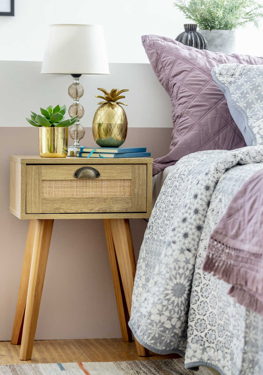 Detalle de un dormitorio con un velador de madera clara con un cajón de ratán. Sobre él hay una lámpara de vidrio con pantalla de tela blanca, un adorno de piña dorada, un macetero dorado con una suculenta artificial. Al costado hay una cama de tonos lilas
