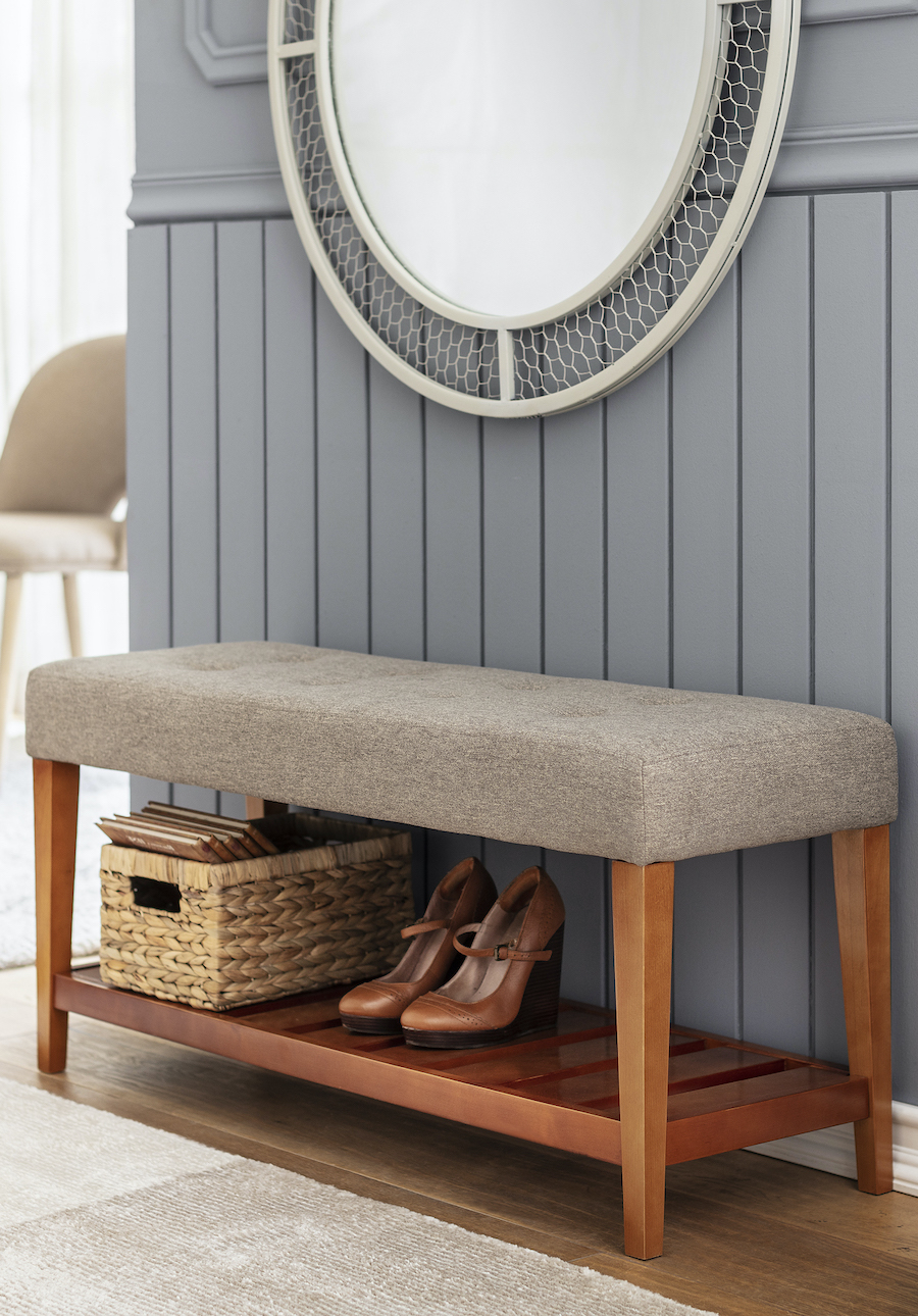 Pasillo estrecho ordenado, con banqueta gris. Bajo ella hay un canasto de mimbre y zapatos cafés. El muro tiene un revestimiento de madera gris y un gran espejo redondo