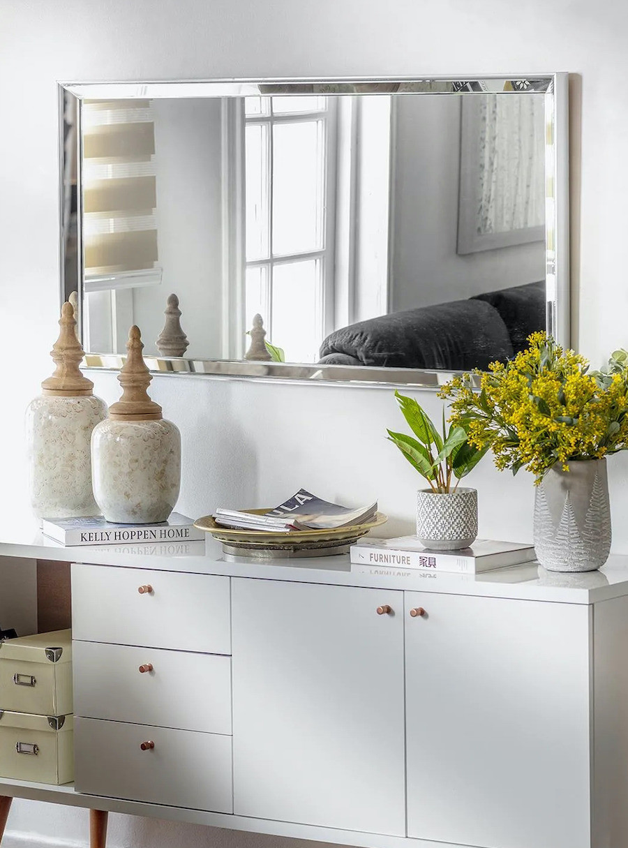 Recibidor con un arrimo blanco y un espejo con doble bisel en el muro. Sobre el arrimos hay floreros, libros y adornos en tonos neutros.