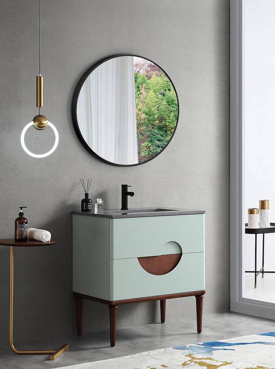 Baño moderno de muros de concreto con un gran espejo redondo de marco negro sobre un vanitorio color verde menta con patas de madera oscura,. La grifería es negra y al costado del espejo cuelga una lámpara dorada.