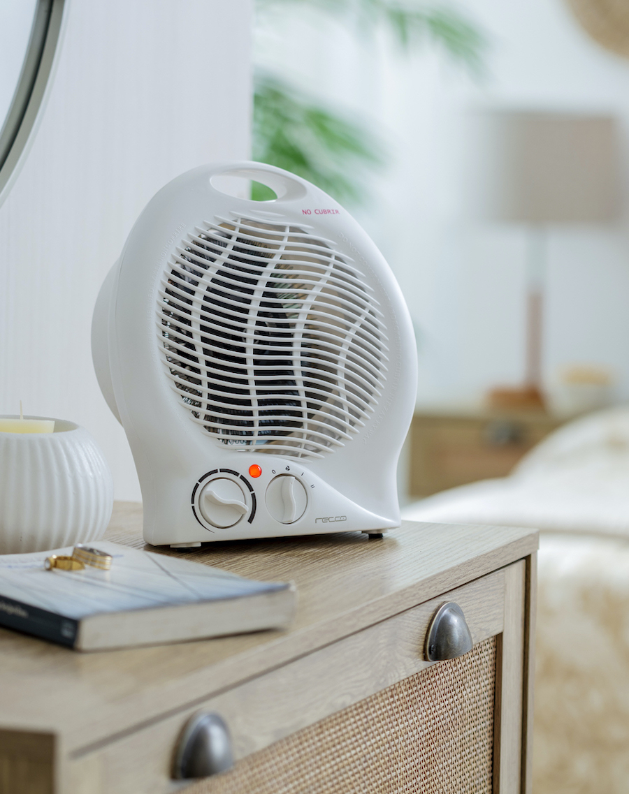 Termoventilador eléctrico blanco, pequeño, disponible en Sodimac. Está ubicado sobre un velador de madera, junto a una vela y un libro.