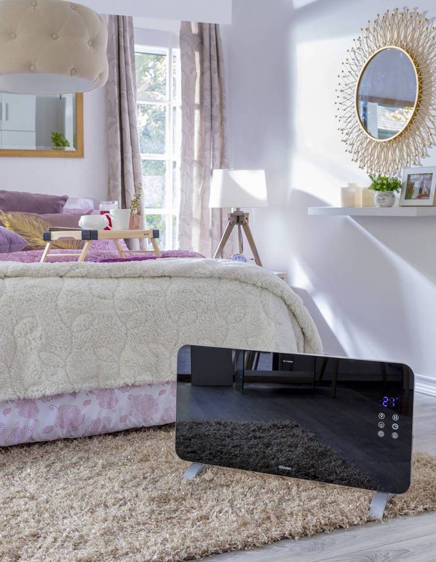 Panel convector de vidrio templado, de color negro y estilo moderno, disponible en Sodimac. Está ubicado en un dormitorio de estilo clásico, sobre una alfombra beige, frente a una cama con plumón rosado y piecera beige