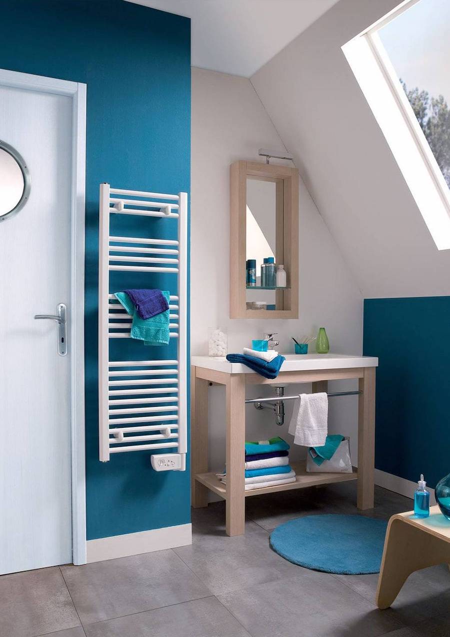 Seca-toallas eléctrico, de pared y de color blanco, disponible en Sodimac. Está colgado en la pared de un baño y sobre él hay dos pequeñas toallas. Las paredes del baño son azules y blancas, con un vanitorio de madera.
