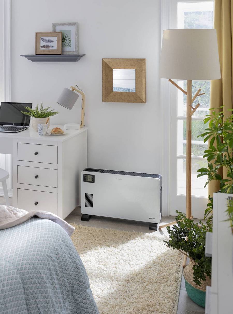 Convector eléctrico disponible en Sodimac. Es de color blanco con patas negras. Ubicado en un dormitorio de estilo moderno, junto a un escritorio blanco con cajoneras y una lámpara de pie