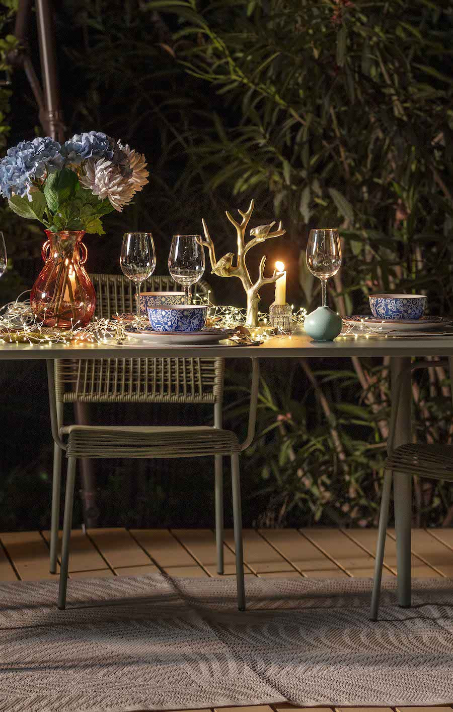 Comedor de terraza de noche. Hay una mesa metálica verde con sillas en el mismo tono. Sobre la mesa hay copas, un florero con hortensias, platos, bowls y velas encendidas.