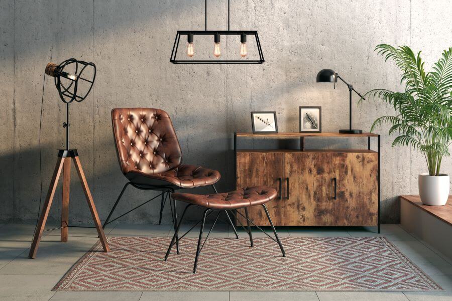 Bodegón industrial con lámpara de techo metálica con tres ampolletas a la vista. Lámpara de pie estilo foco, con patas de madera y pantalla metálica abierta. Mueble de madera con lámpara de mesa de escritorio negra. Sitial de cuero con apoya pies.