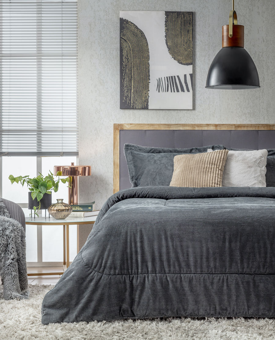 Dormitorio estilo ejecutivo urbano. Cama de dos plazas con plumón gris. Cojines beige y blanco. Respaldo de cama gris con marco de madera. Lámpara de techo metálica negra.