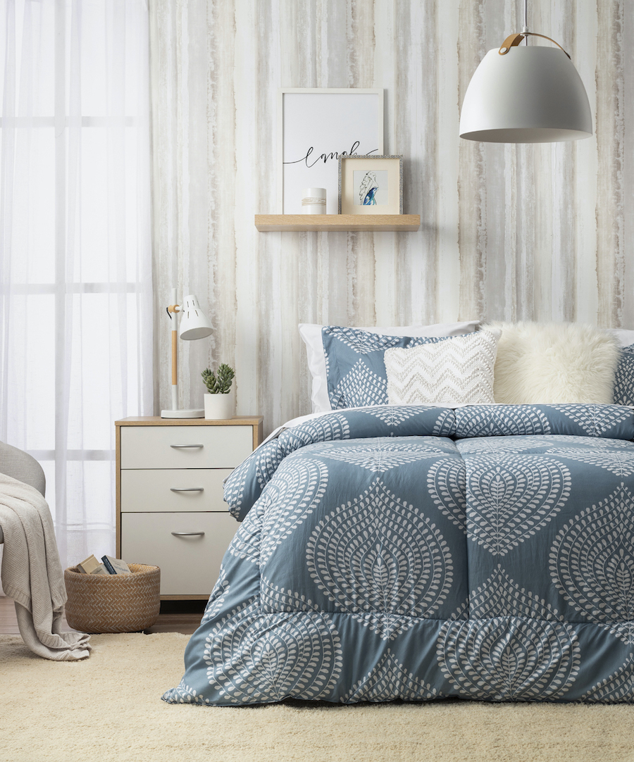 Dormitorio de estilo nórdico. Cama de dos plazas con plumón celeste con figuras blancas. Cojines de color blanco. Sobre la cama hay una repisa flotante de madera con adornos. Velador estilo moderno con cajones blancos.