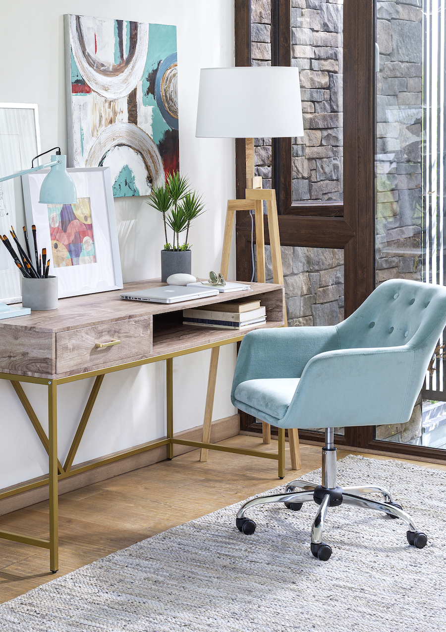 Escritorio de madera con patas doradas, silla celeste de terciopelo y una lámpara de pie de estilo nórdico. Sobre el escritorio hay cuadros de colores.