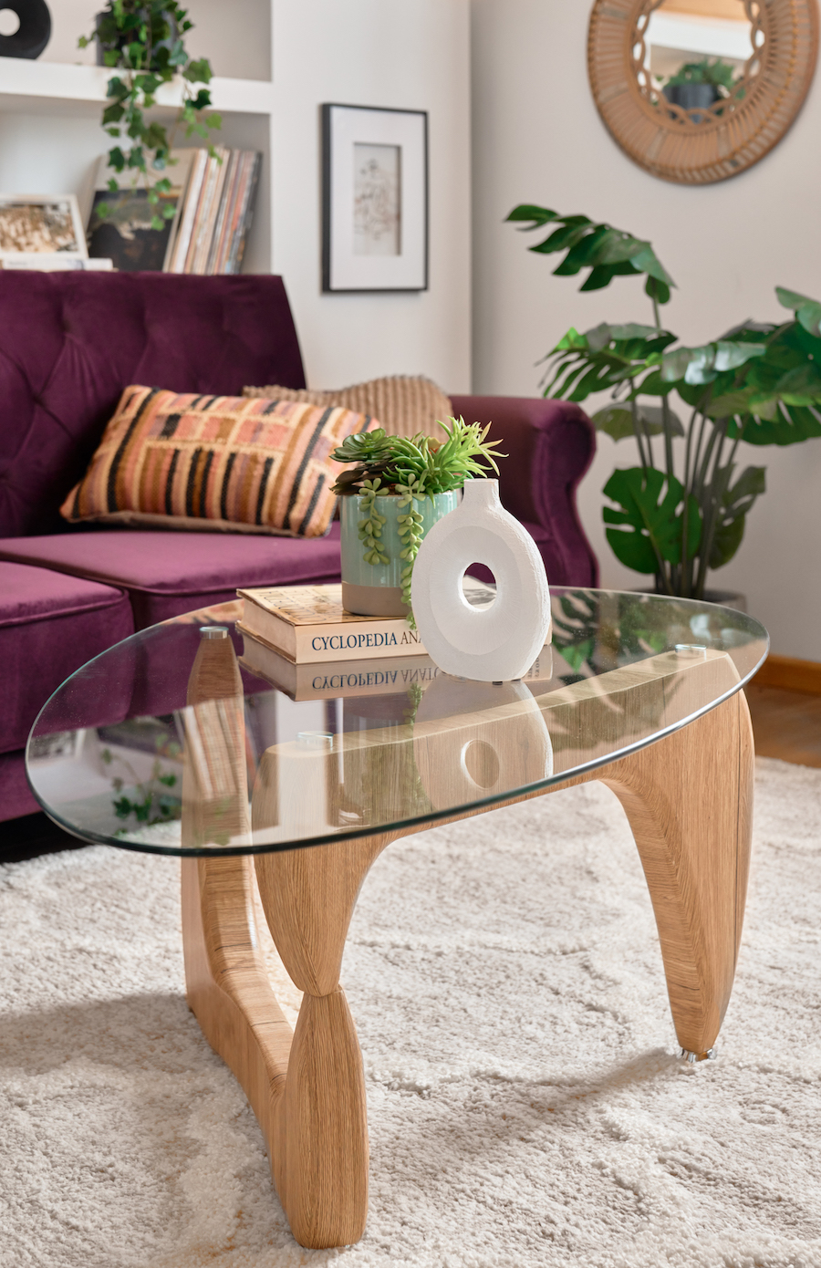 Detalle de una mesa de centro de vidrio con diseño irregular y base de madera. Sobre ella hay un florero blanco y una suculenta sobre un libro. De fondo hay un sofá morado junto a una planta monstera artificial.