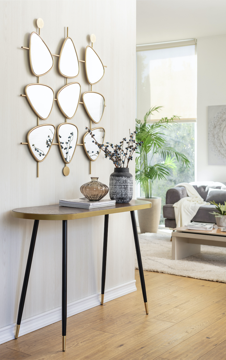 Entrada con un arrimo de patas delgadas y negras y cubierta en forma ovalada con detalles dorados. Sobre el arrimo hay un florero con ramas, un jarro de vidrio y una pila de revistas de decoración. En la pared hay un espejo escultórico con pequeños espejos