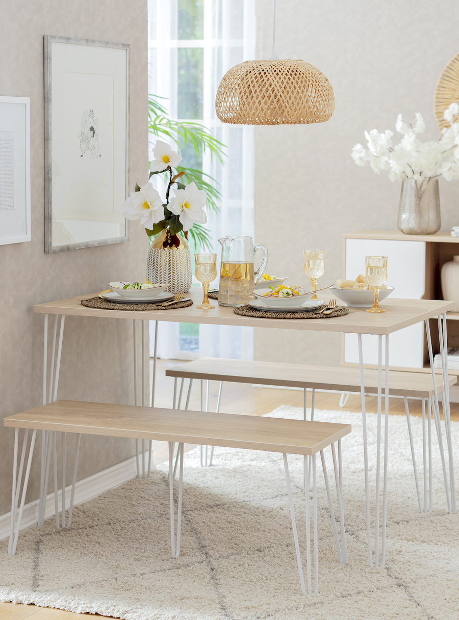 Comedor de colores claros con un juego de comedor de madera clara con patas blancas delgadas y 2 bancas que funcionan como asientos. Sobre la mesa hay menaje en tonos crema y grises, así como un florero de vidrio con flores blancas en su interior.