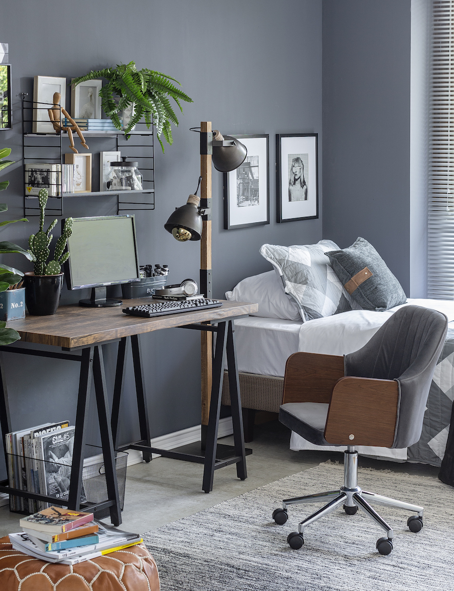 Dormitorio de estilo industrial con una cama con cojines y cobertor grises, paredes grises y un escritorio de patas metálicas negras y cubierta de madera oscura.