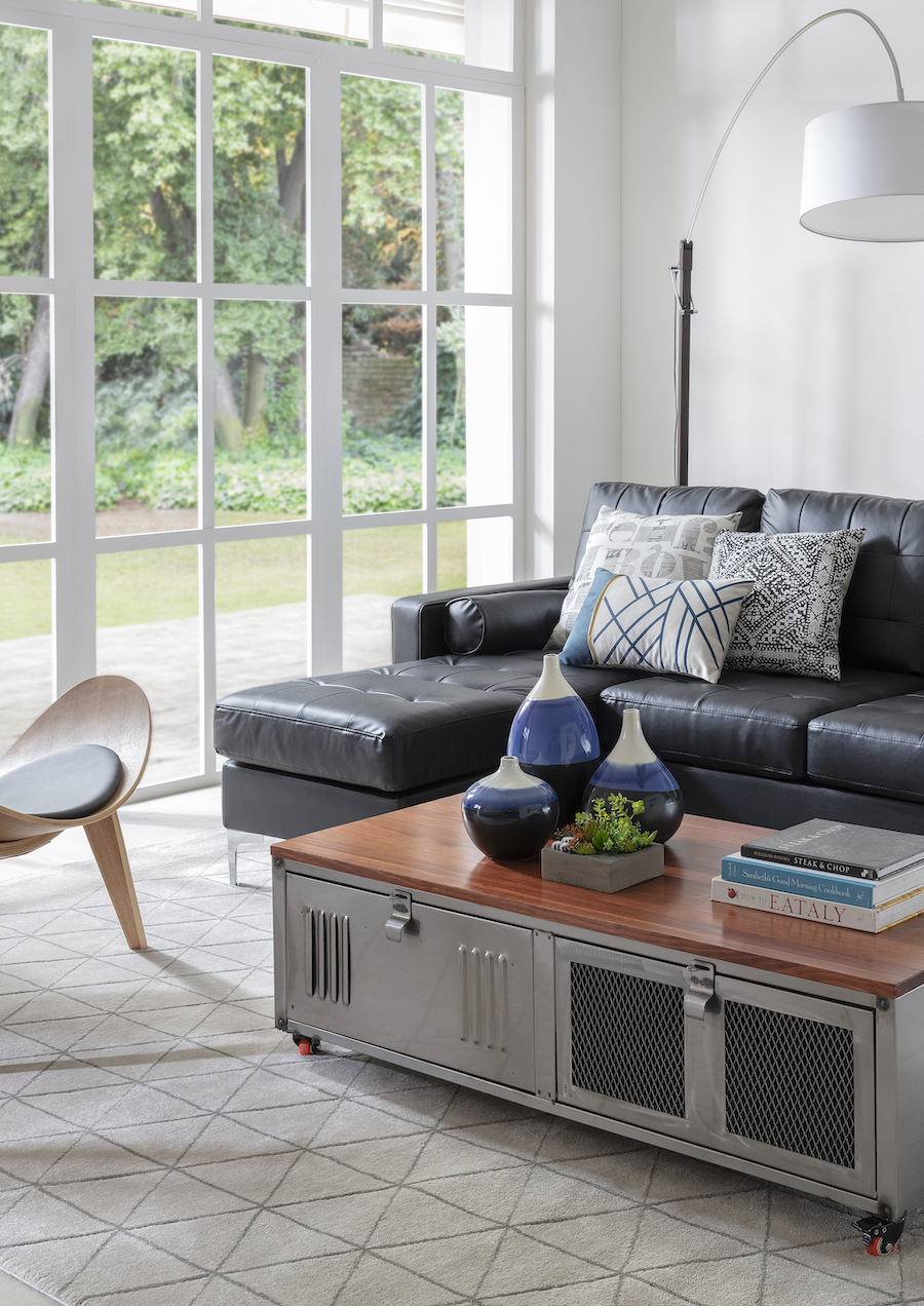 Sala de estar de estilo ejecutivo urbano con un sofá seccional negro con cojines grises, una mesa de centro industrial gris con cubierta de madera y al fondo, hay un gran ventanal sin cortinas, aportando a la modernidad del espacio.