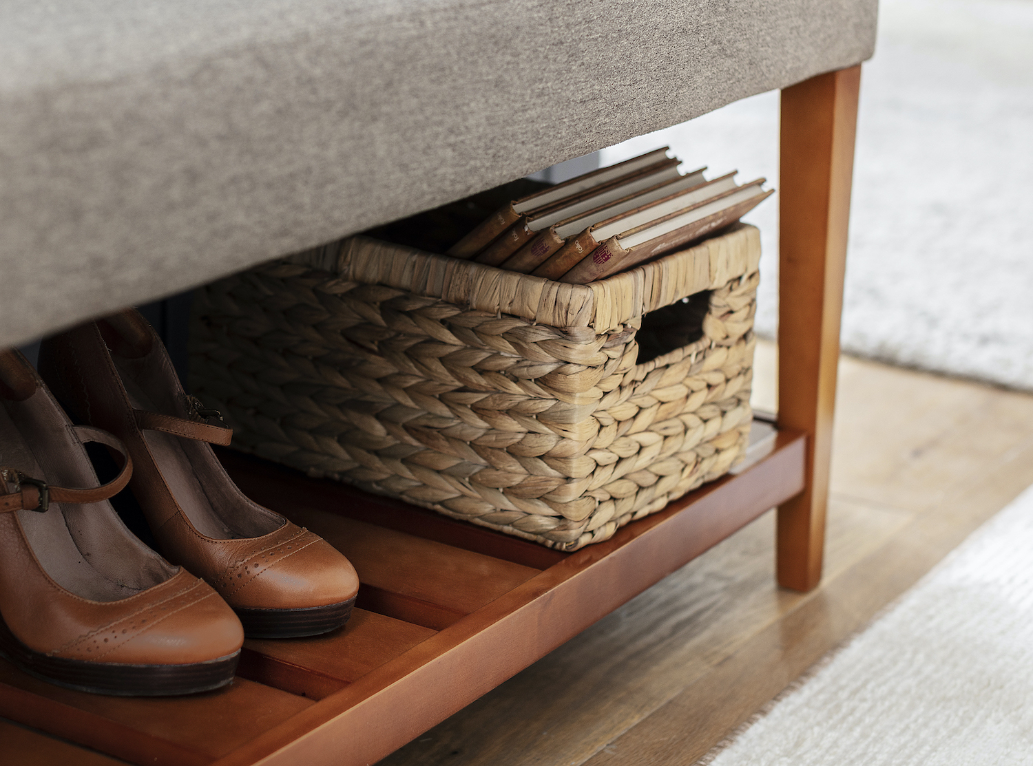 Detalle de una banqueta de madera, en cuya repisa inferior hay un par de zapatos y una caja de fibras naturales con libros en su interior.