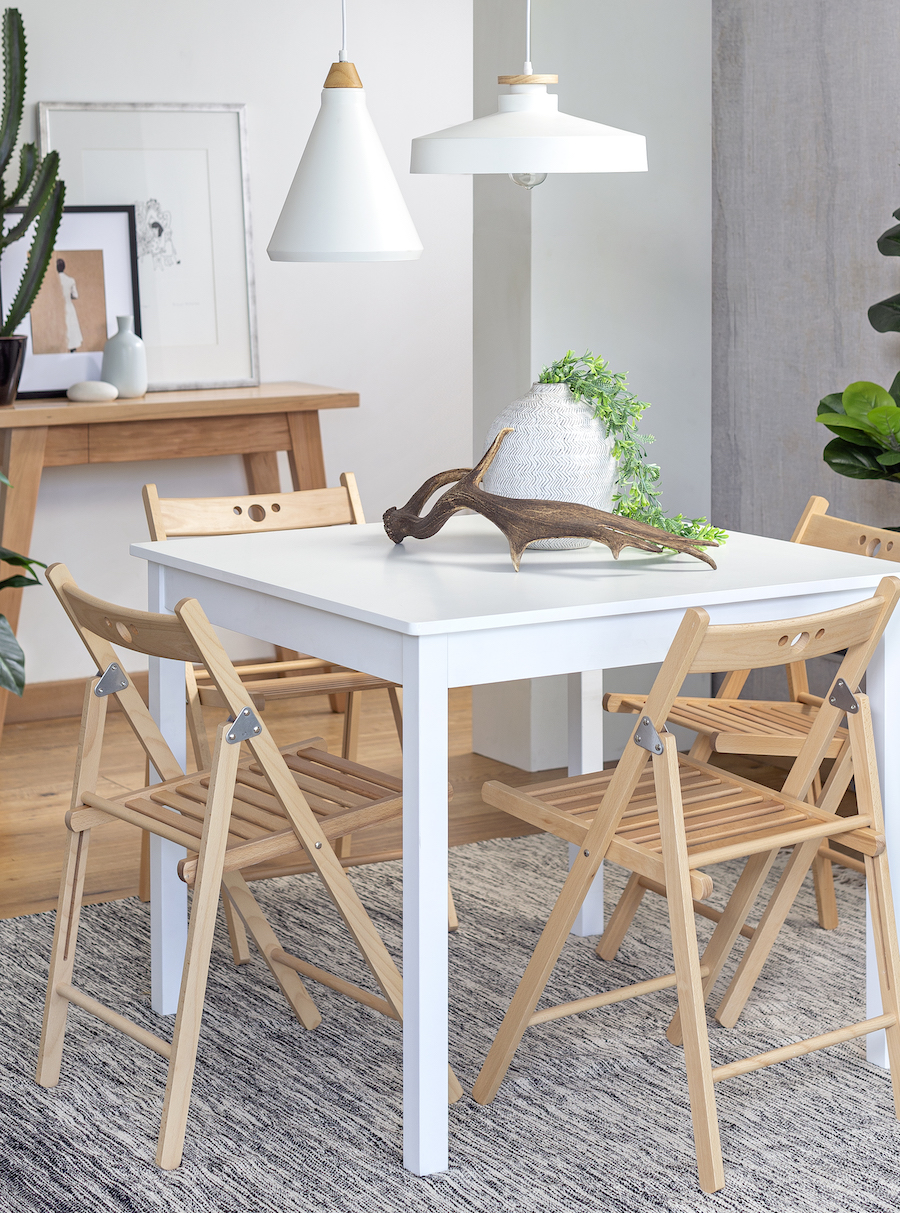 Comedor de estilo nórdico con una mesa rectangular blanca, 4 sillas plegables de madera clara, una lámpara colgante blanca y adornos de mesa inspirados en la naturaleza. En el piso hay una alfombra gris.