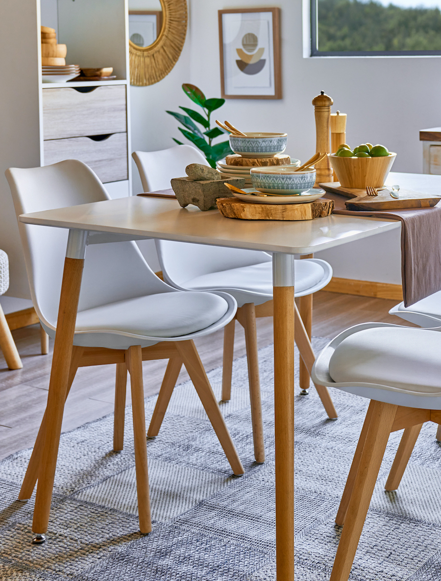 Comedor pequeño con una mesa de comedor de patas de madera y cubierta blanca y 4 sillas estilo eames con patas de madera. Sobre la mesa hay menaje de madera.