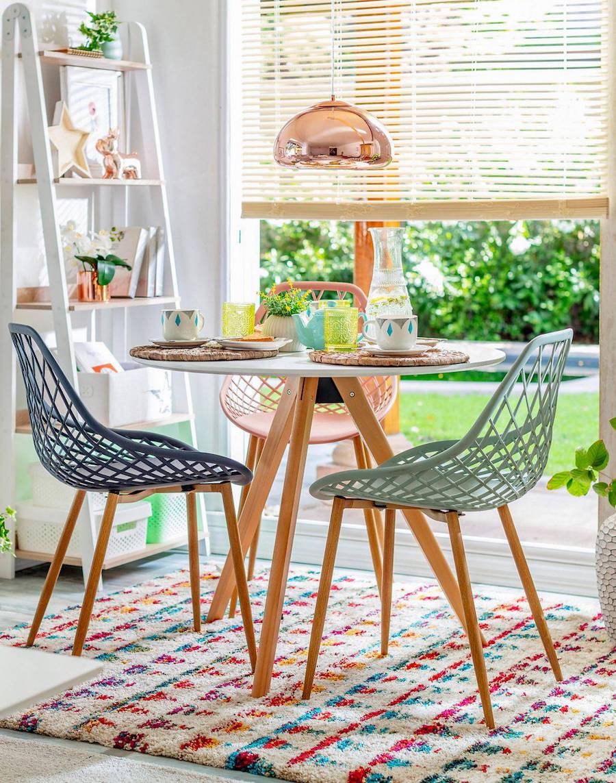 Comedor pequeño con una mesa redonda blanca con patas de madera, sillas de color pastel con patas de madera clara. En el piso hay una alfombra de colores estilo shaggy y desde el techo cuelga una lámpara con acabado de bronce.