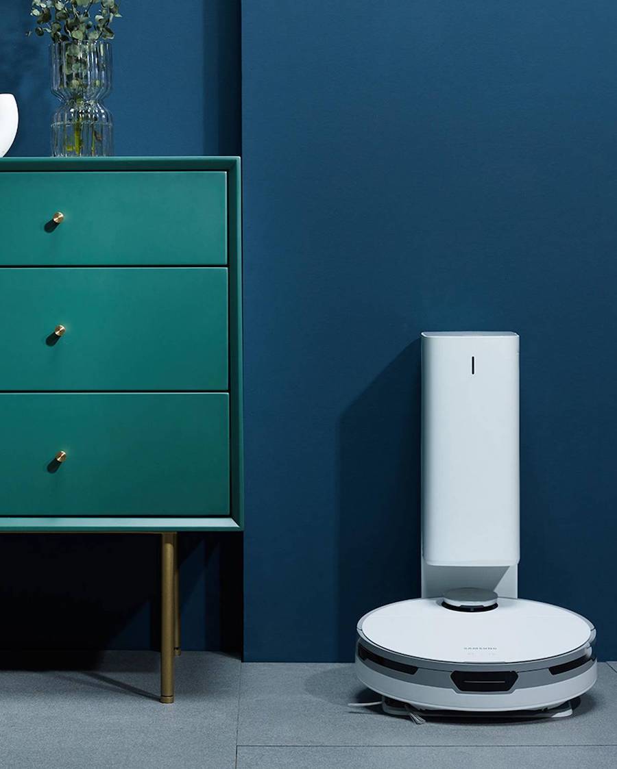 Detalle de una cómoda verde azulada junto a una aspiradora robot blanca.