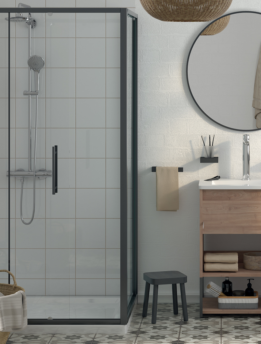 Baño de estilo industrial con muros blancos, ducha con mampara de bordes negros y cerámica blanca en la pared. Junto a ella hay un mueble vanitorio de madera con patas negras y sobre él, un gran espejo redondo con marco negro.