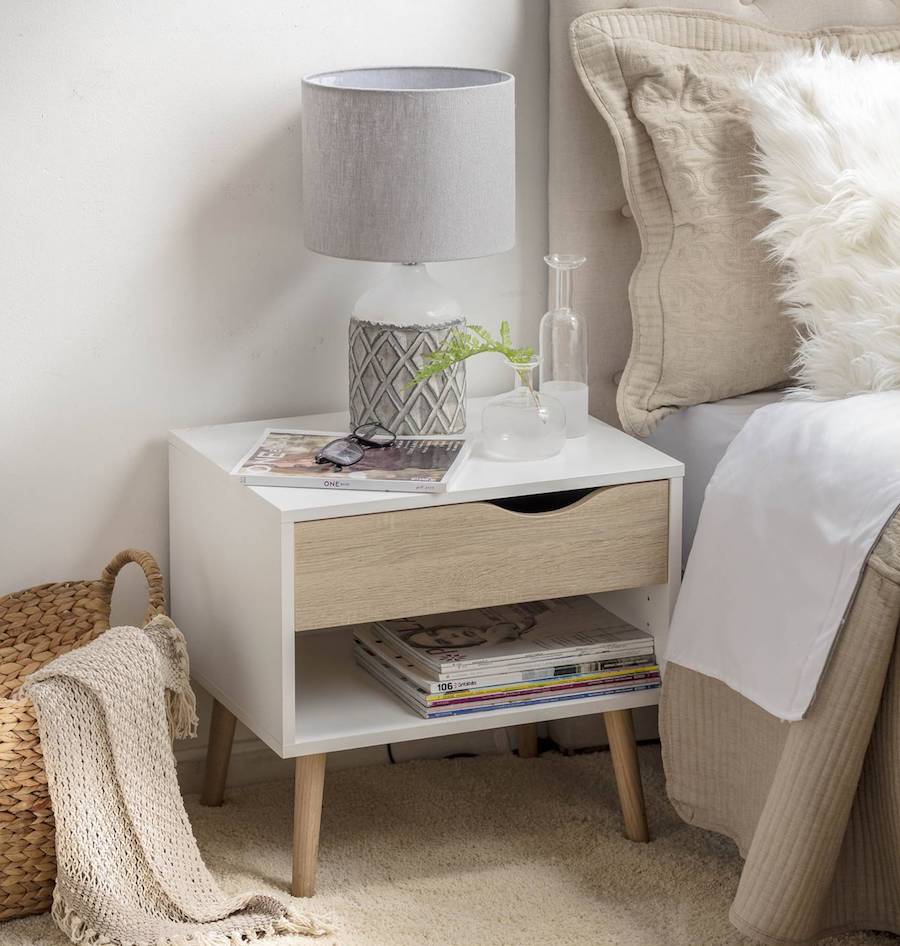 Velador de estilo nórdico junto a una cama con textiles de color neutro. Sobre él hay una lámpara, un libro y adornos de vidrio.