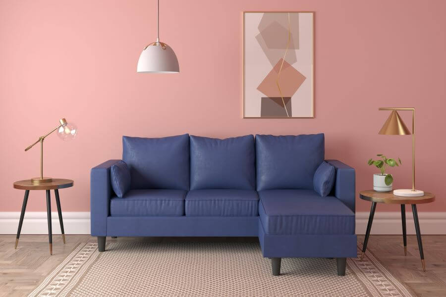 Bodegón con sillón en L azul. Lámpara de techo con pantalla blanca curva. Dos mesas laterales, de madera. En una hay una lámpara de mesa con pie dorado y gran ampolleta redonda a la vista. En la otra hay una lámpara de base blanca, pie y pantalla dorada