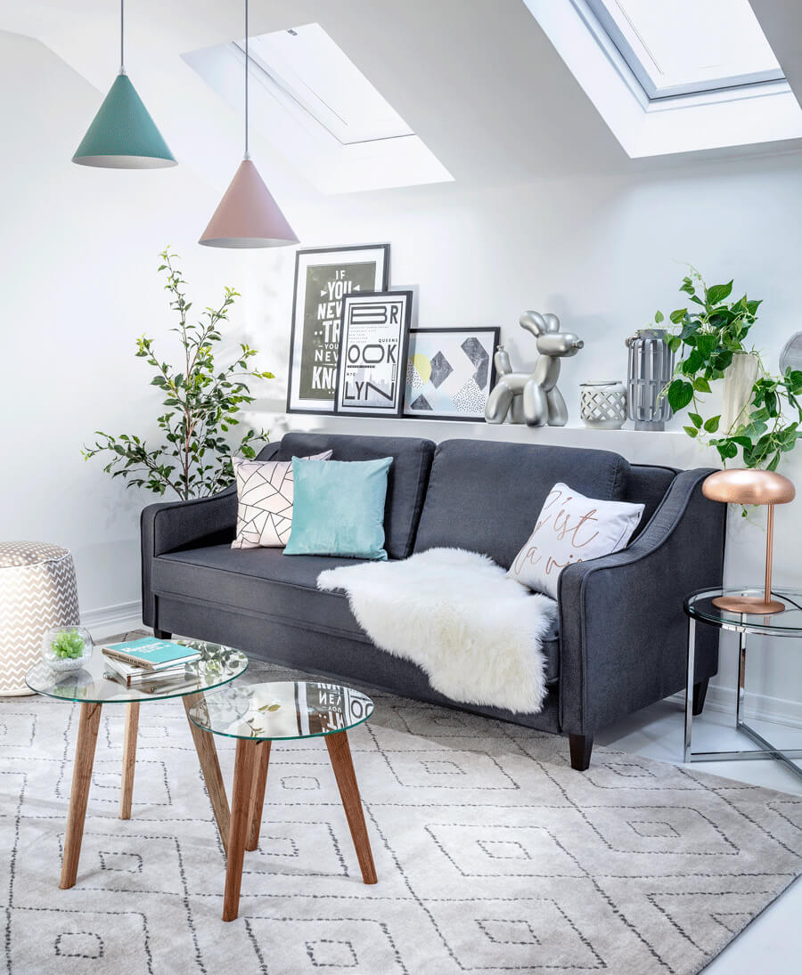 Sillón gris oscuro de dos cuerpos, con cojines estampados en tonos pasteles y manta blanca peluda. Desde el techo cuelgan dos lámparas con pantalla cónica, una es rosada pastel y la otra, turquesa. La alfombra es blanco invierno con líneas grises, sobre ella se ubican dos mesitas de centro redondas con cubierta de vidrio circular.