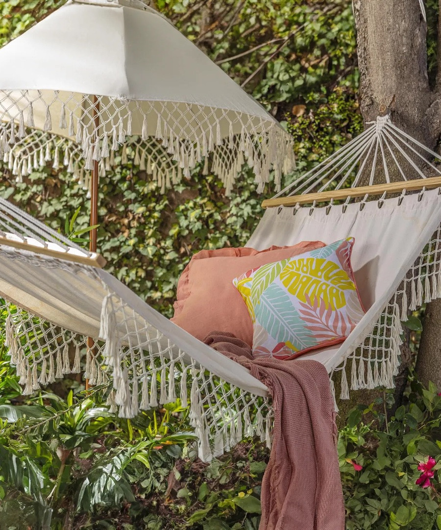 Hamaca blanca con flecos junto a un quitasol blanco con flechos. Sobre una hamaca hay cojines y una manta rosada.