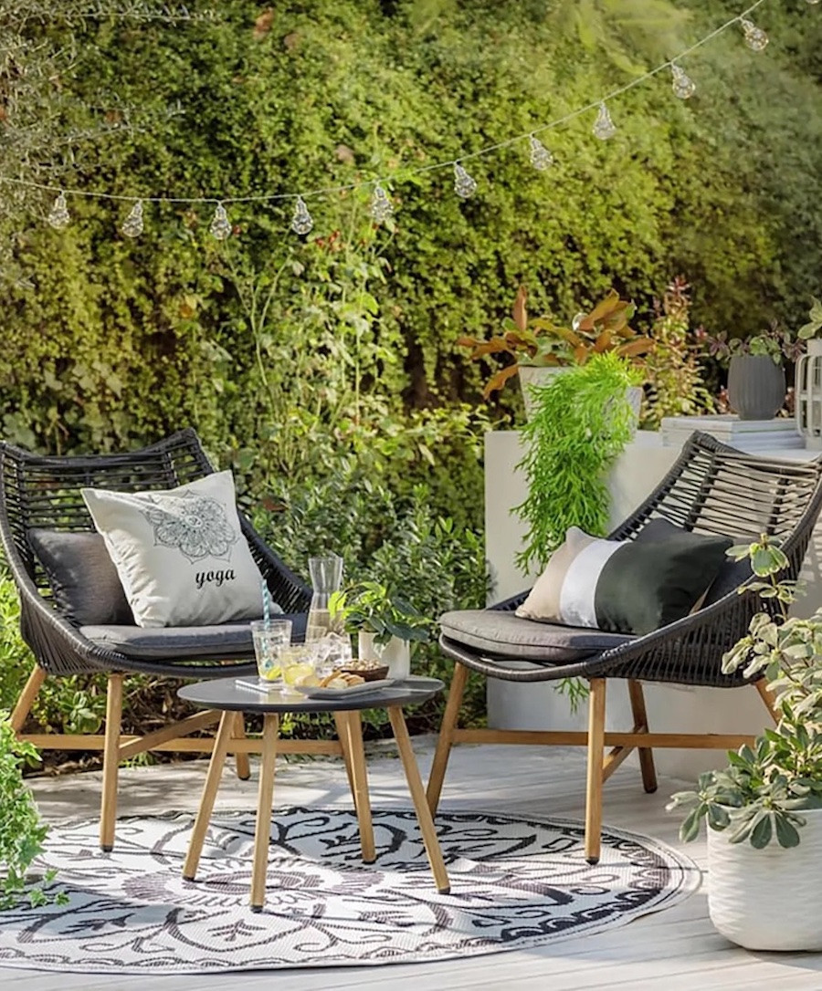 Juego de living de terraza para dos personas con 2 sofás y una mesa redonda auxiliar. Los asientos son trenzados y de color gris oscuro, mientras que las patas son de madera clara. Sobre ellos hay cojines en tonos neutros. 