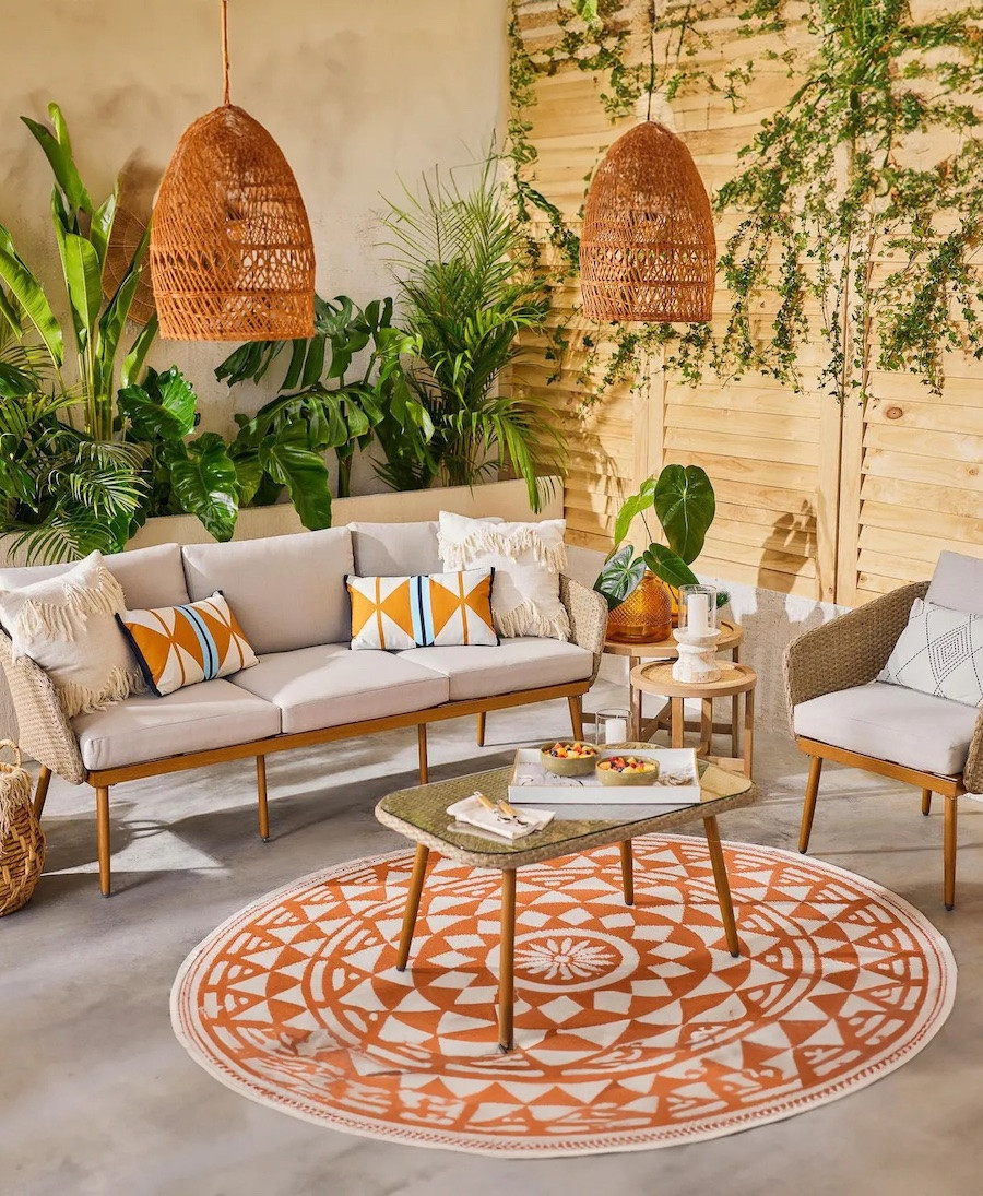 Juego de living de terraza estilo Boho chic. Hay un sofá de madera con cojines blancos y amarillos, un sillón para una persona y una mesa de centro sobre una alfombra redonda con estampado tribal anaranjado. Desde el techo cuelgan 2 lámparas de mimbre.