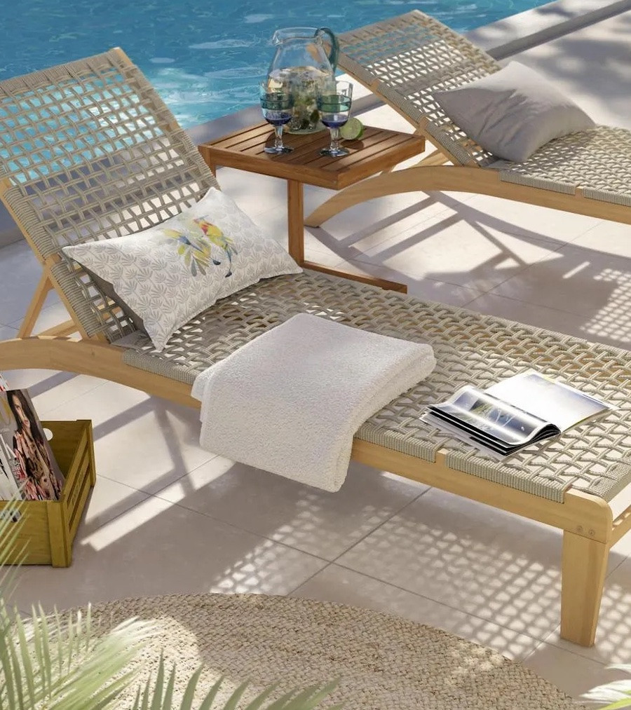 Reposera de terraza de madera y tejido gris.Está al lado de una piscina y sobre ella hay un cojín blanco con un estampado, una toalla y una revista.