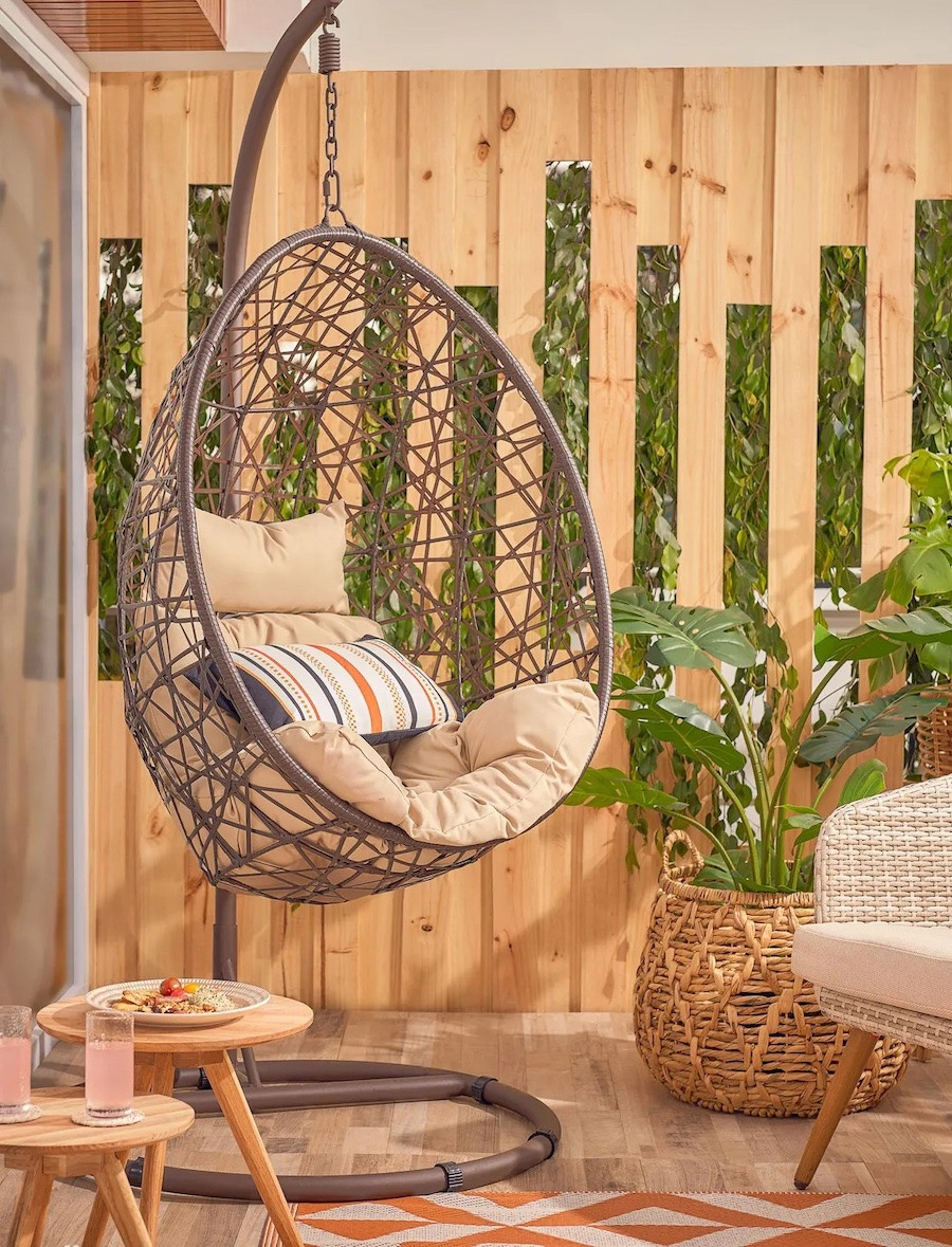 Silla colgante de terraza estilo nido café, tiene un asiento color beige, un cojín rayado y está al lado de una mesa auxiliar redonda y una gran planta dentro de un canasto de mimbre.