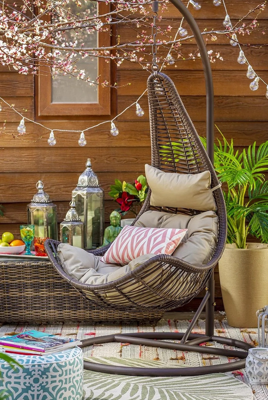 Silla colgante para terraza con un asiento café claro y un cojín con estampado blanco y rosado. De fondo hay plantas, una guirnalda de luces, faroles y vasos.