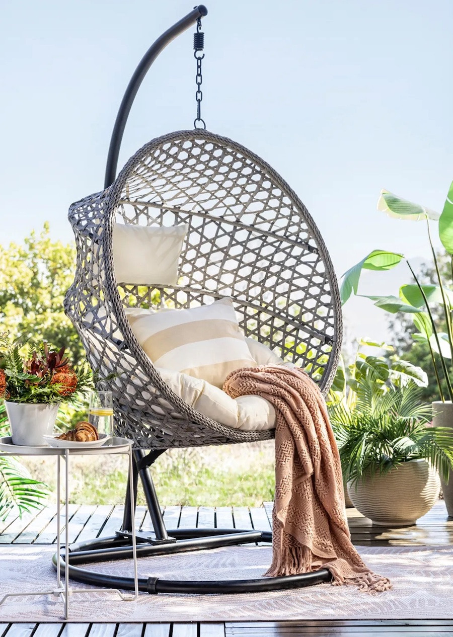 Silla colgante para terraza gris en forma de nido con asiento y cojines en blanco crema. Sobre ellos hay una manta palo rosa. Alrededor hay muchas plantas de distintos tamaños.